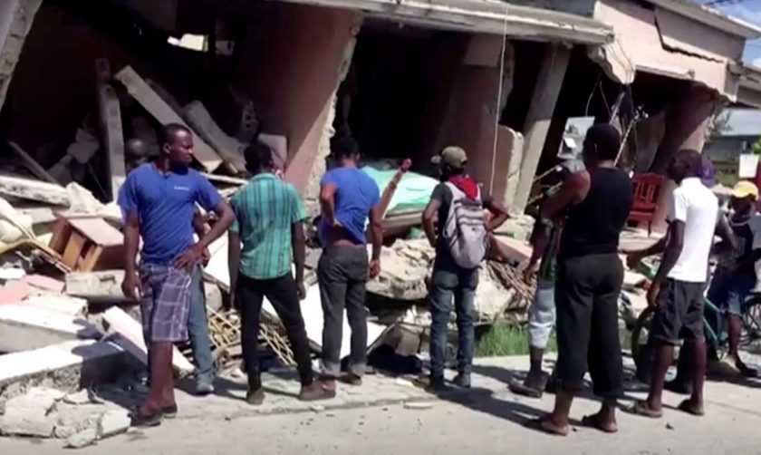 People stand in front of a collapsed building following an earthquake, in Les Cayes, Haiti, in this still image taken from a video obtained by Reuters on August 14, 2021.   REUTERS TV via REUTERS THIS IMAGE HAS BEEN SUPPLIED BY A THIRD PARTY. MANDATORY CREDIT.